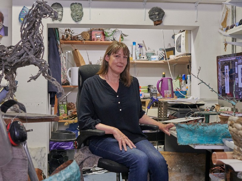 An artist sits in her studio. She looks at the camera and is dressed casually in a long sleeve blue shirt and jeans. She is surrounded by artists equipment and sculptures.